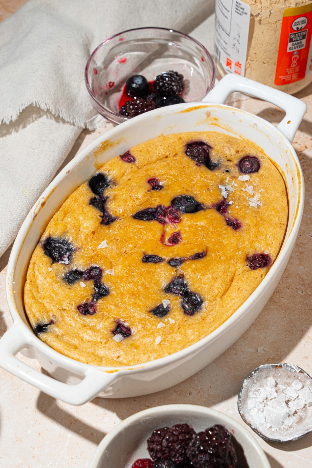 A baked protein pancake bowl with blueberries. The bowl is on a tan counter with a white cloth napkin and small bowl of fruit.