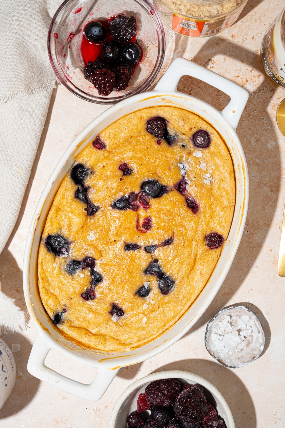 A baked protein pancake bowl with blueberries. The bowl is on a tan counter with a white cloth napkin and small bowl of fruit.