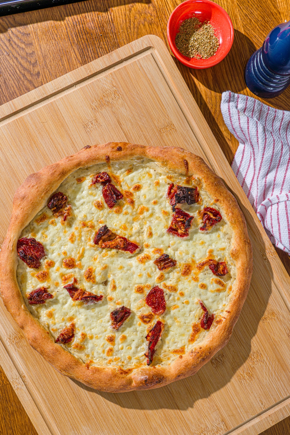 A wooden board with a whole white pizza topped with roasted peppers. The board is on a wooden counter with a white striped napkin.
