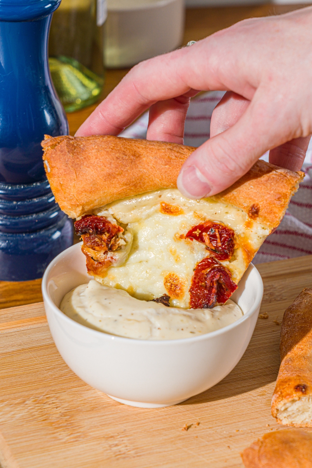 A hand dipping a slice of white pizza with roasted peppers into a bowl of garlic white pizza sauce. The bowl is on a wooden board with additional slices of pizza.