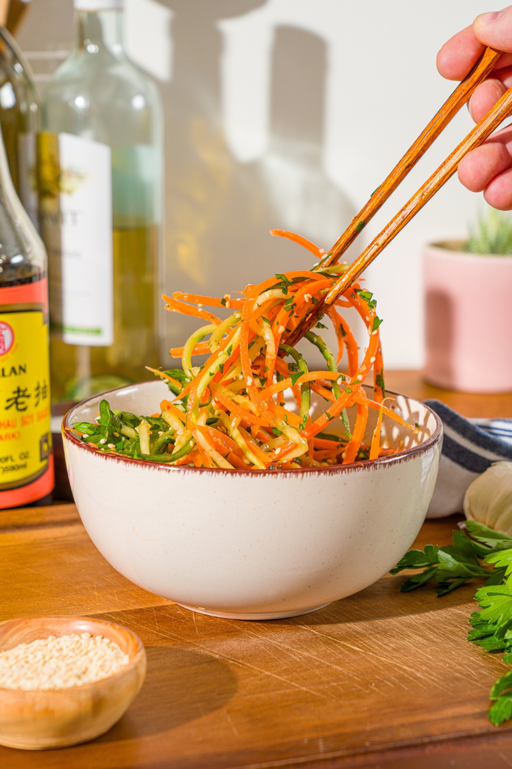 A white bowl with carrot cucumber salad tossed in dressing. A pair of chopsticks is taking a bite from the salad. The bowl is on a wooden board.