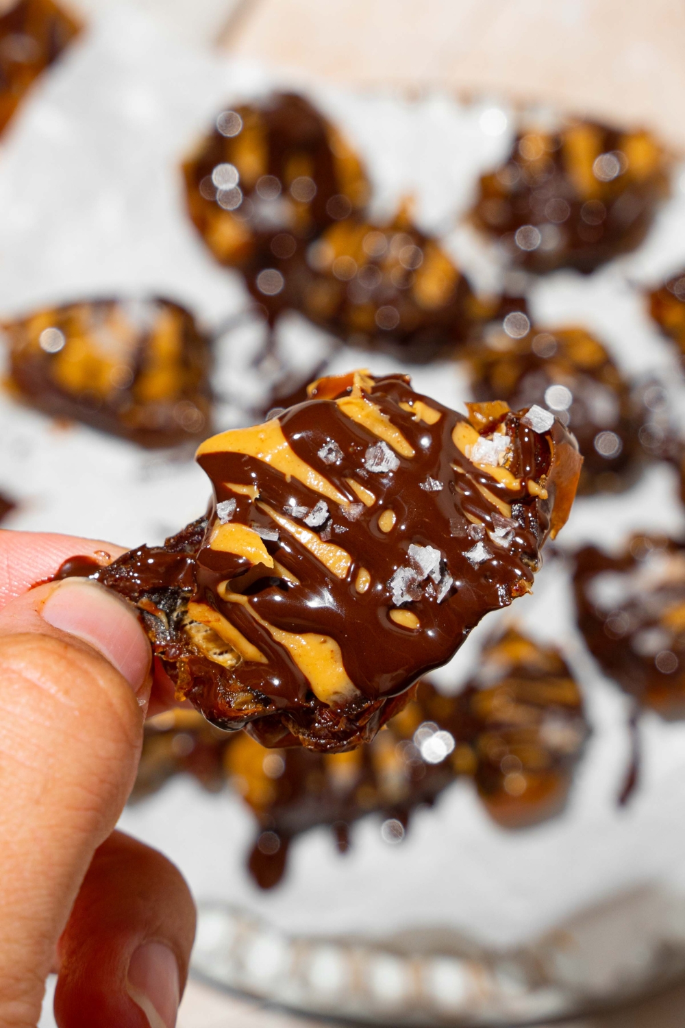 A close up of a hand holding a date stuffed with peanut butter and topped with chocolate and sea salt. There is a plate of peanut butter dates in the background.