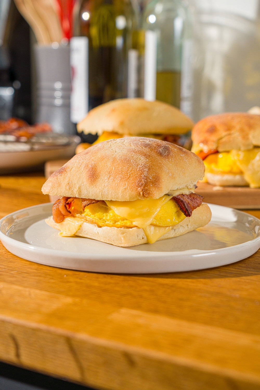 A white plate with a Starbucks gouda bacon egg sandwich. The plate is on a wooden counter with a wooden board with additional sandwiches.