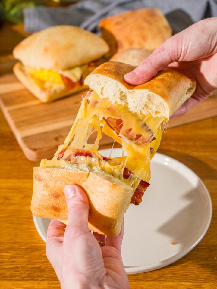 A close up of a hand splitting a cheesy Starbucks gouda bacon sandwich in half. There is a wooden board with additional sandwiches blurred in the background.