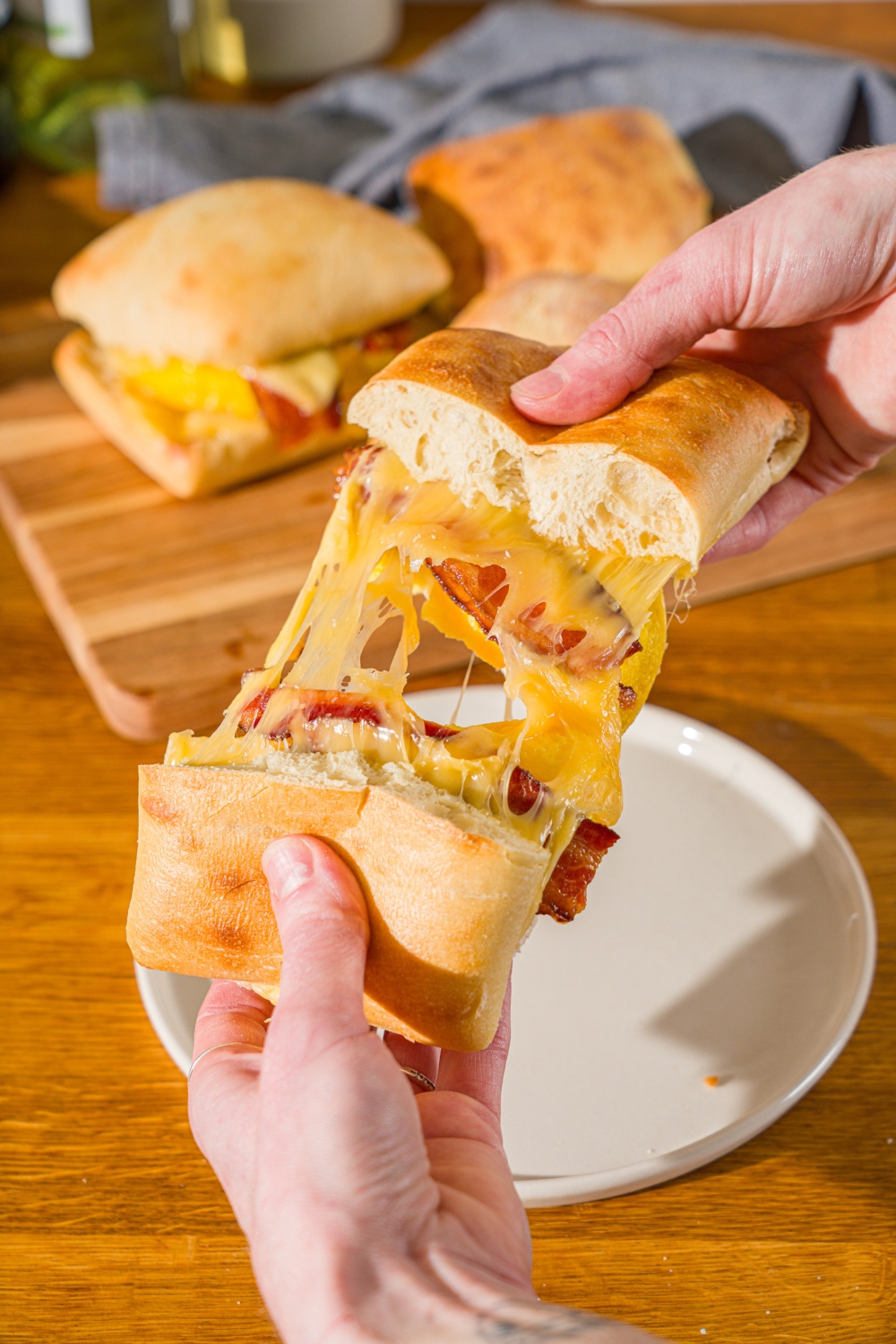 A close up of a hand splitting a cheesy Starbucks gouda bacon sandwich in half. There is a wooden board with additional sandwiches blurred in the background.