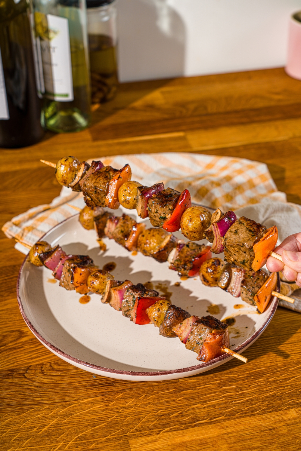 A white plate with three grilled steak kabobs with marinated steak, peppers, onions, and potatoes. A hand is holding one of the kabobs. The plate is on a wooden counter with a yellow striped napkin.