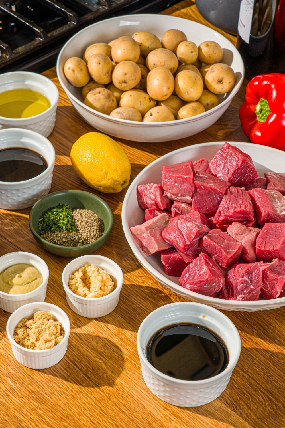 A wooden counter with several bowls in various sizes containing ingredients to make steak kabob marinade including steak, potatoes, soy sauce, olive oil, garlic, lemon, Dijon musard, brown sugar, and seasonings.