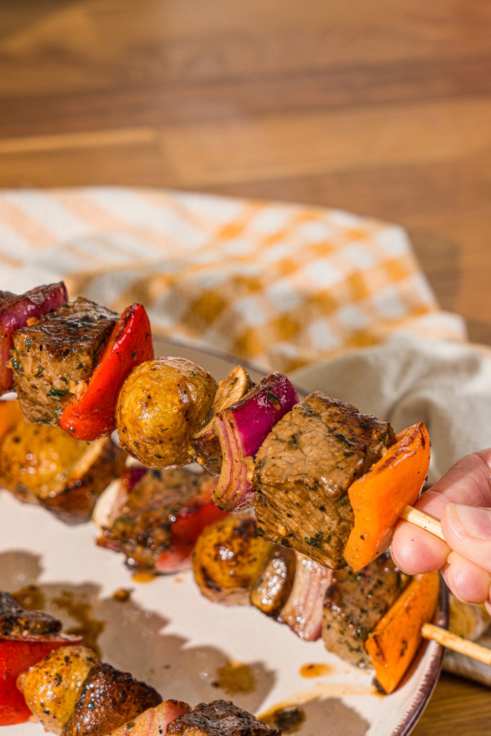 A hand holding a steak kabob with marinated steak, peppers, onion, and potatoes. There is a plate of kabobs on a wooden counter blurred in the background.
