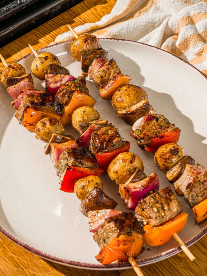 A white plate with three grilled steak kabobs with marinated steak, peppers, onions, and potatoes. The plate is on a wooden counter with a yellow striped napkin.