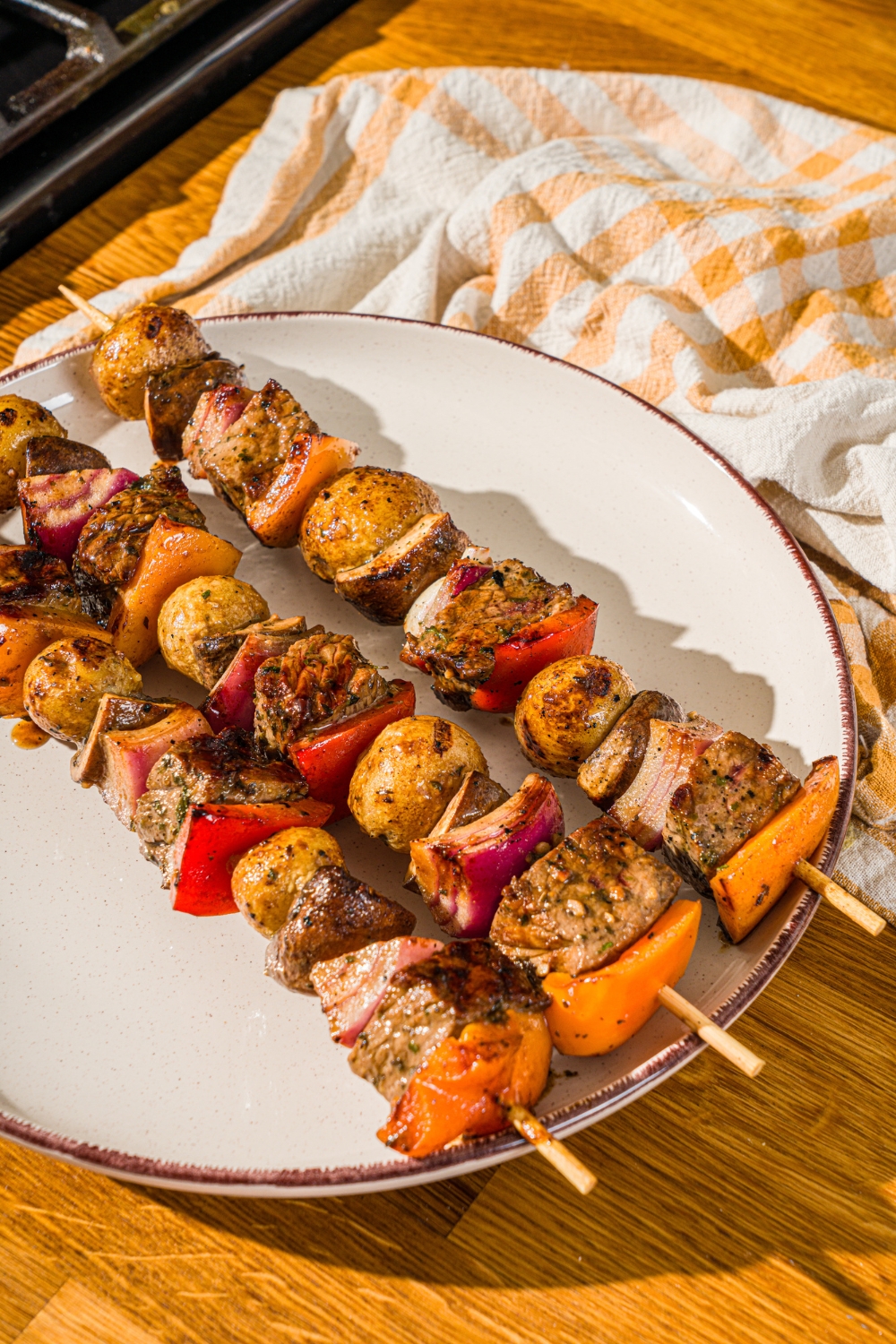 A white plate with three grilled steak kabobs with marinated steak, peppers, onions, and potatoes. The plate is on a wooden counter with a yellow striped napkin.