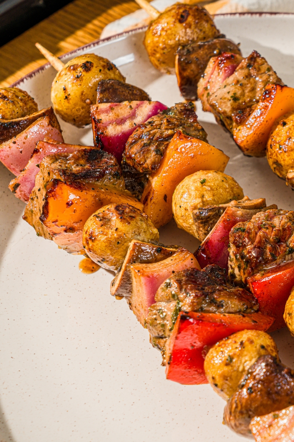 A white plate with three grilled steak kabobs with marinated steak, peppers, onions, and potatoes. The plate is on a wooden counter with a yellow striped napkin.