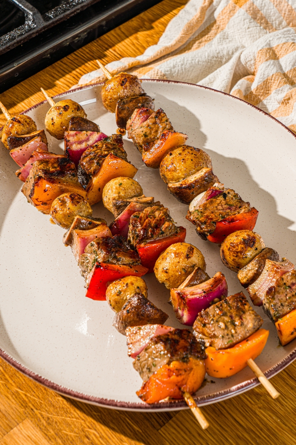 A white plate with three grilled steak kabobs with marinated steak, peppers, onions, and potatoes. The plate is on a wooden counter with a yellow striped napkin.