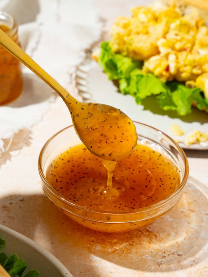 A bowl with sweet onion sauce with a spoon dipping in the sauce. The bowl is on a tan counter with a white plate with an open sandwich and a white cloth napkin.