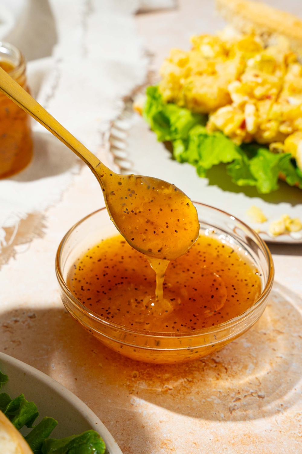 A bowl with sweet onion sauce with a spoon dipping in the sauce. The bowl is on a tan counter with a white plate with an open sandwich and a white cloth napkin.