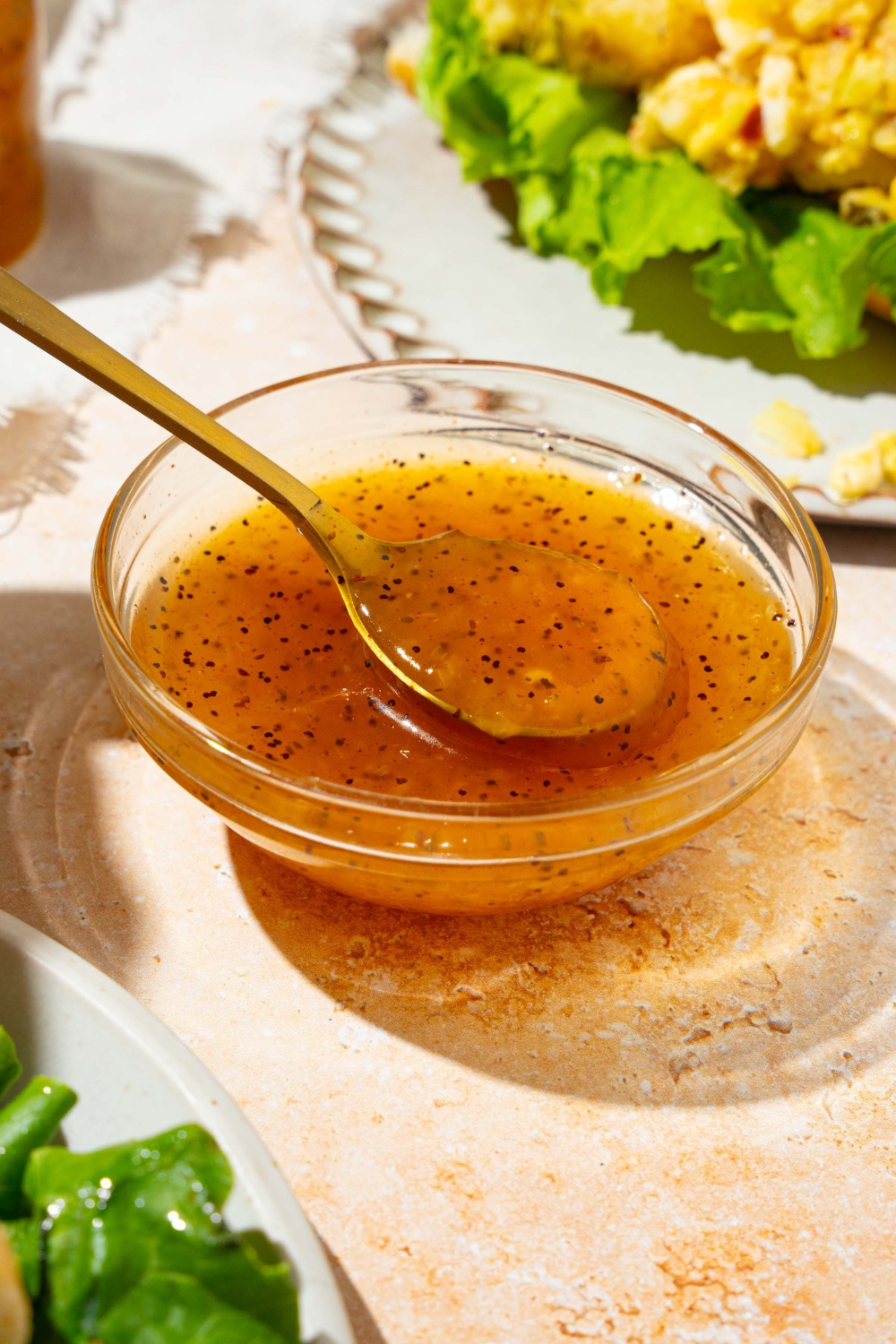 A bowl with sweet onion sauce with a spoon dipping in the sauce. The bowl is on a tan counter with a white plate with an open sandwich and a white cloth napkin.