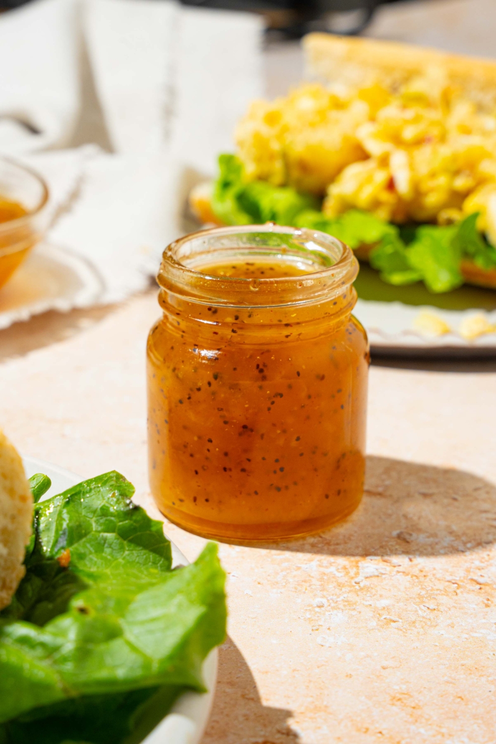 A jar with sweet onion sauce. The jar is on a tan counter with a plate with an open sandwich and a white cloth napkin.