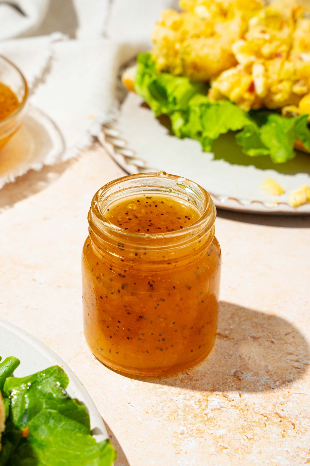 A jar with sweet onion sauce. The jar is on a tan counter with a plate with an open sandwich and a white cloth napkin.