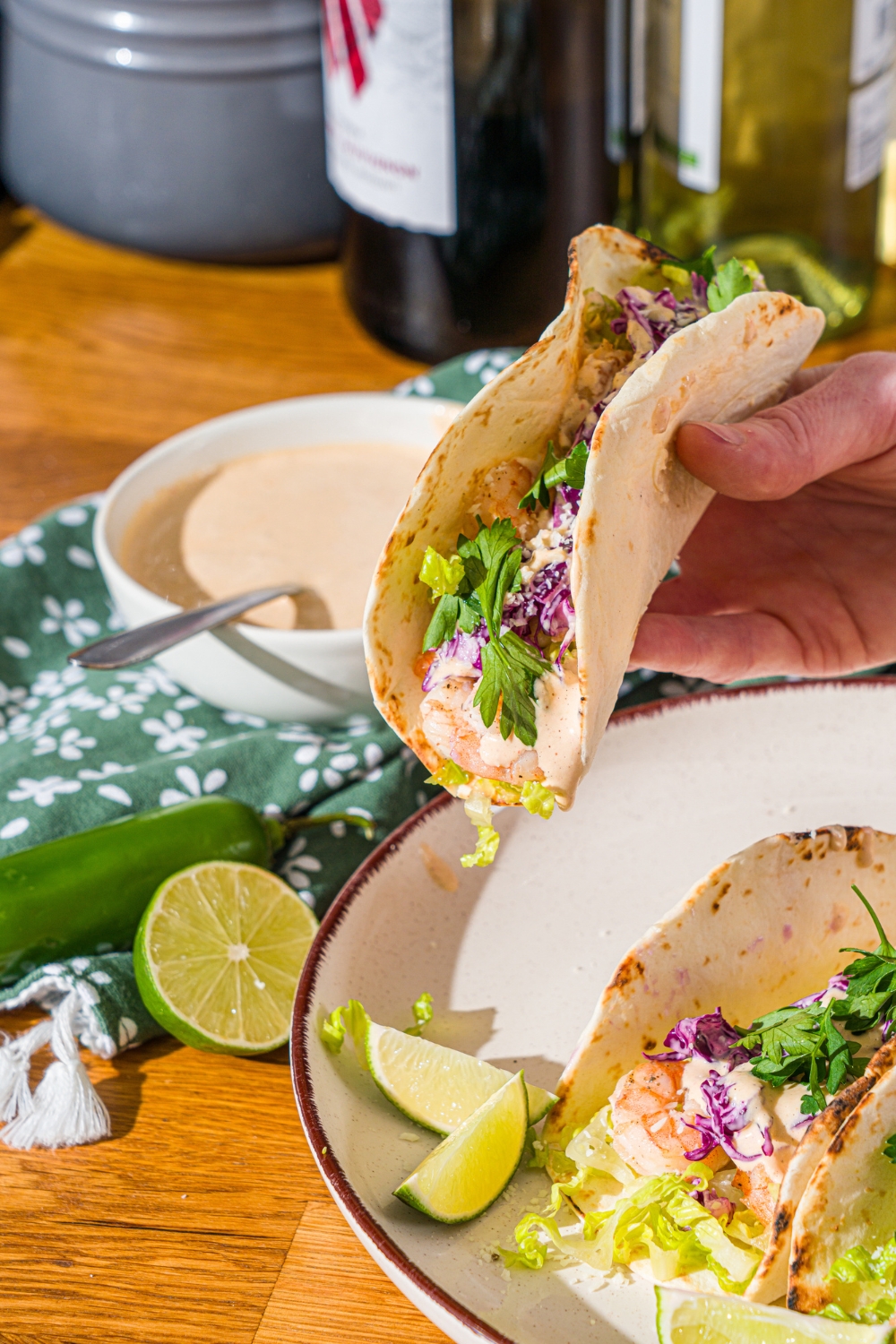 A hand holding a shrimp taco with baja sauce and fresh cilantro over a white plate with shrimp tacos. The plate is on a wooden counter with a bowl of baja sauce and green cloth napkin.