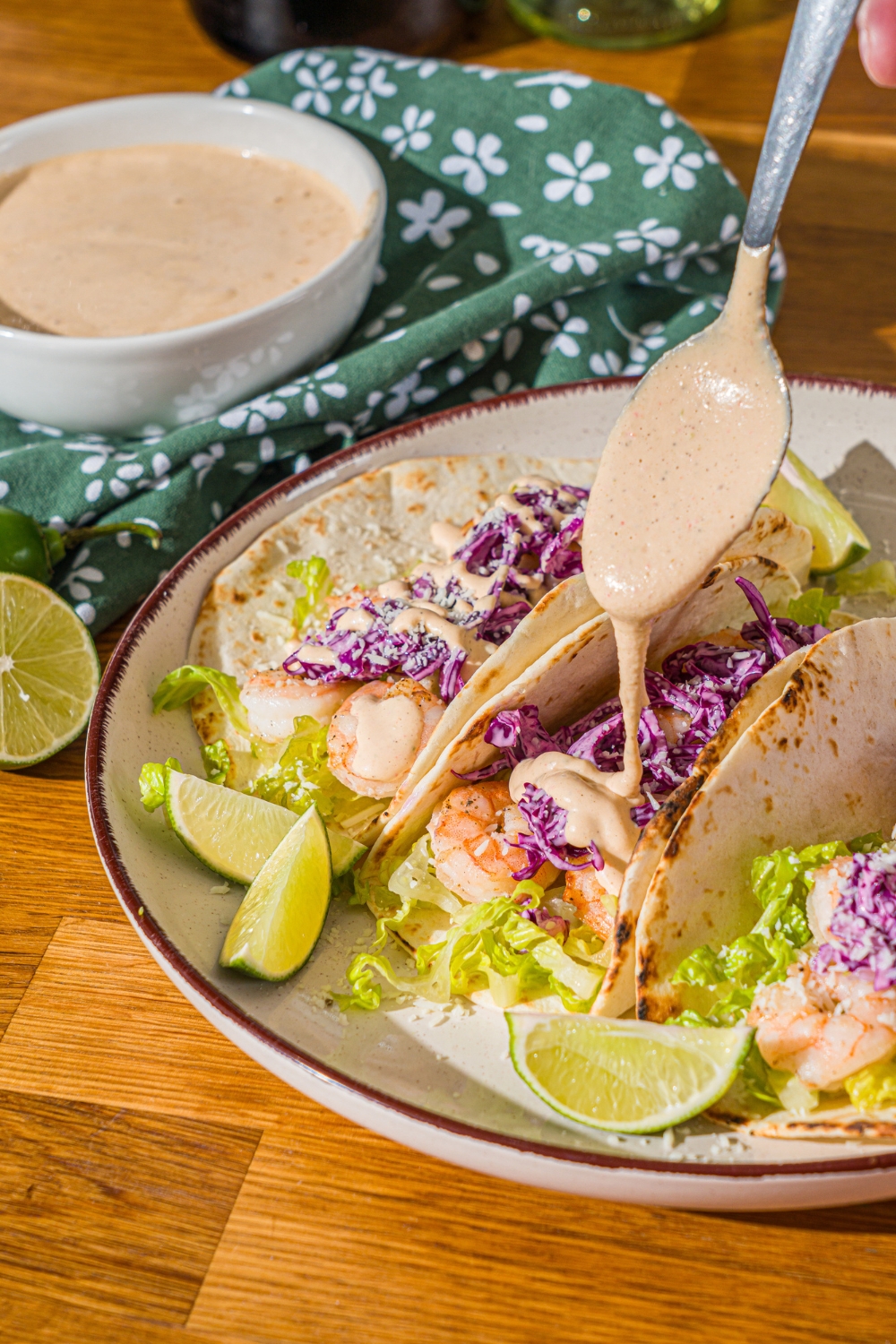 A white plate with shrimp tacos topped with shredded cabbage. A spoon is drizzling baja sauce over the taco. The plate is on a wooden counter with a green cloth napkin.