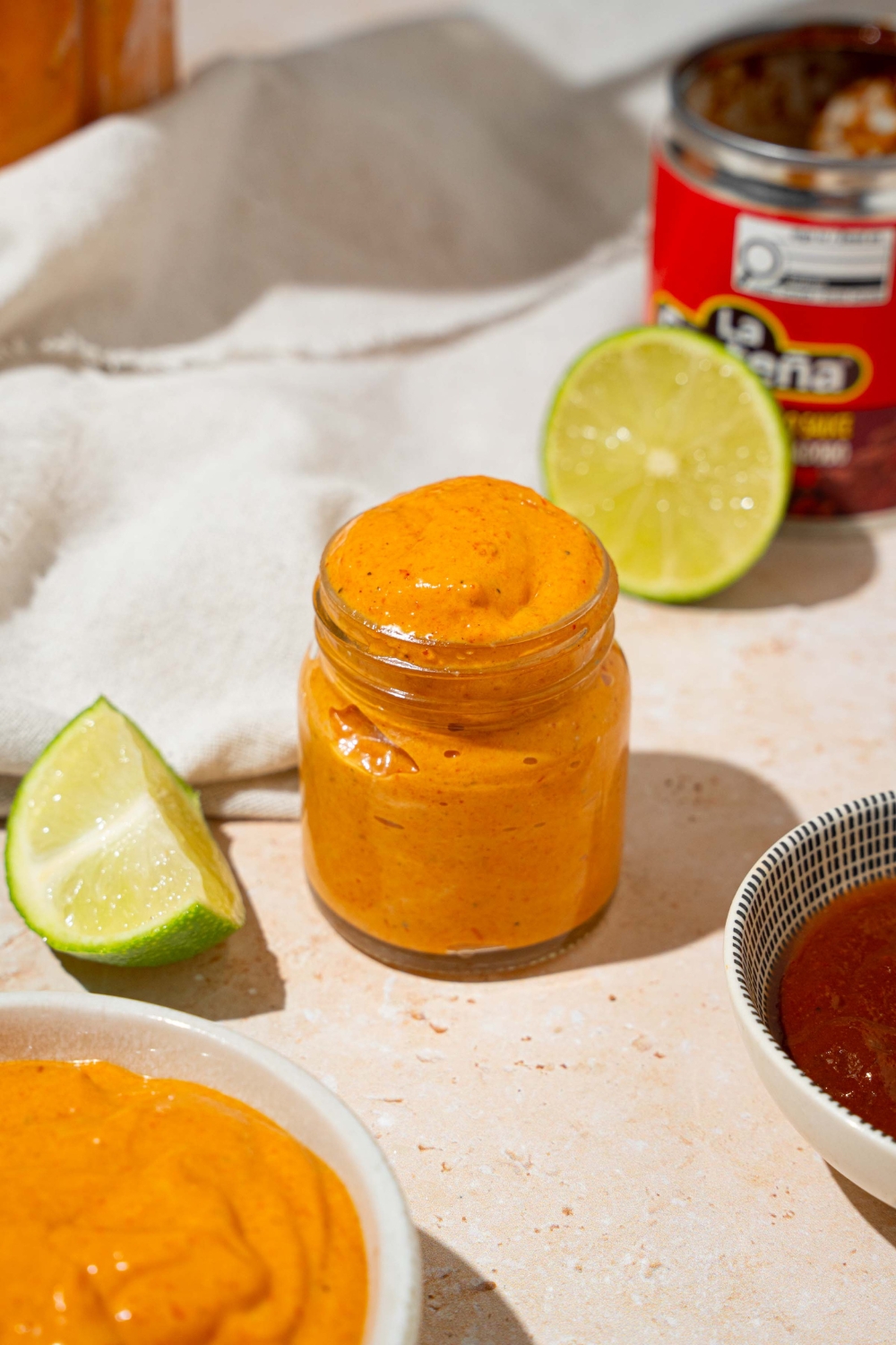 A small glass jar with Taco Bell chipotle sauce. The jar is on a tan counter with a bowl of sauce, lime, and white cloth napkin.