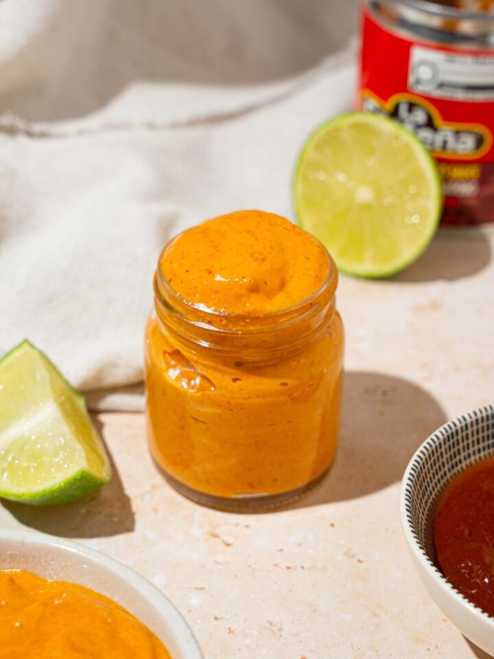 A small glass jar with Taco Bell chipotle sauce. The jar is on a tan counter with a bowl of sauce, lime, and white cloth napkin.