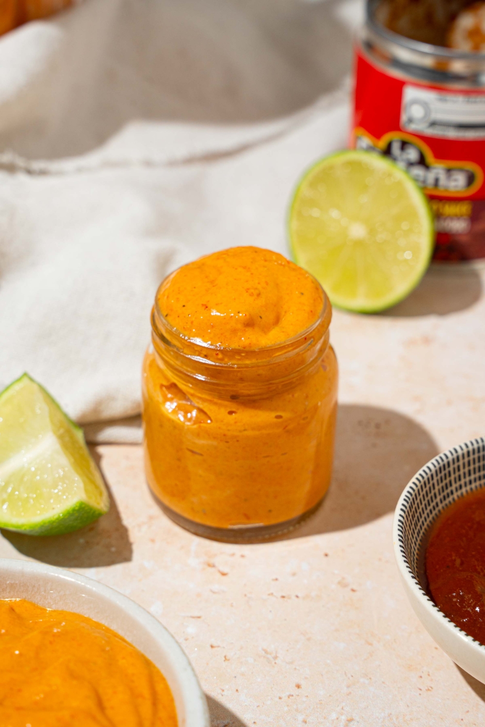 A small glass jar with Taco Bell chipotle sauce. The jar is on a tan counter with a bowl of sauce, lime, and white cloth napkin.