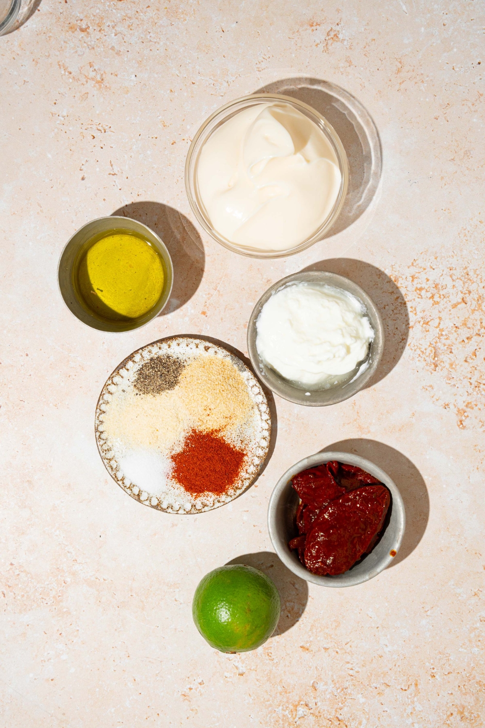 A tan counter with several bowls in various sizes containing ingredients to make Taco Bell chipotle sauce including mayo, sour cream, chipotle peppers, lime, and seasonings.