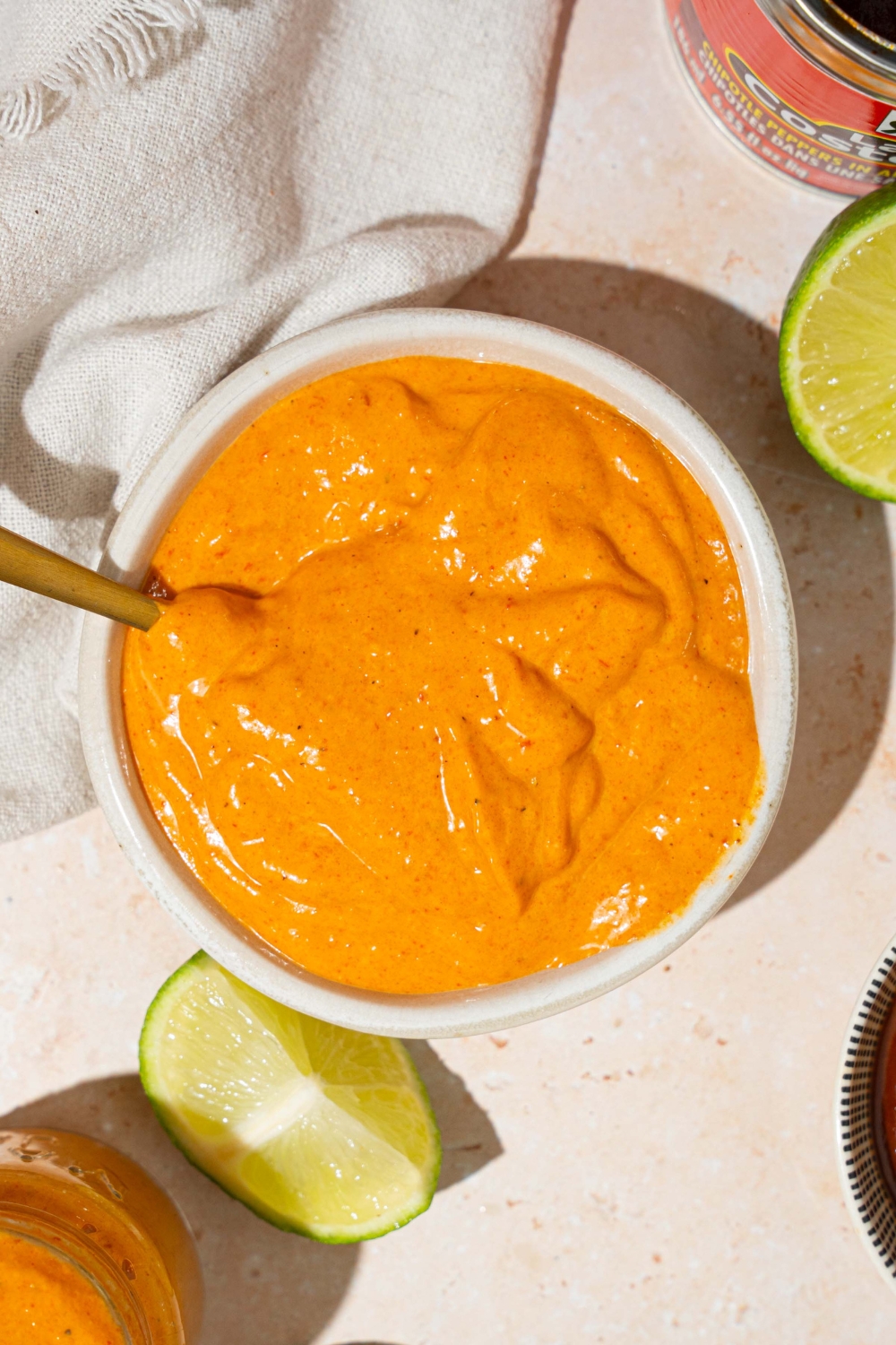 A white bowl with Taco Bell chipotle sauce with a spoon in the bowl. The bowl is on a tan counter with lime and a white cloth napkin.