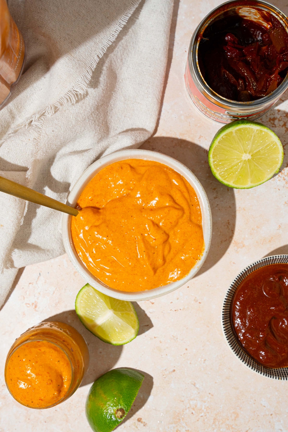 A white bowl with Taco Bell chipotle sauce with a spoon in the bowl. The bowl is on a tan counter with a jar of sauce, lime, and a white cloth napkin.