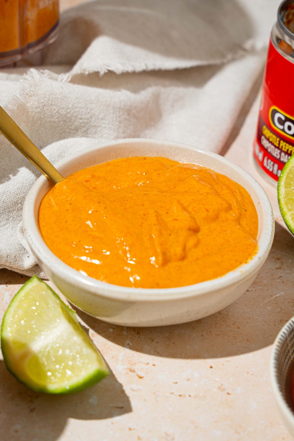 A white bowl with Taco Bell chipotle sauce with a spoon in the bowl. The bowl is on a tan counter with lime, and a white cloth napkin.