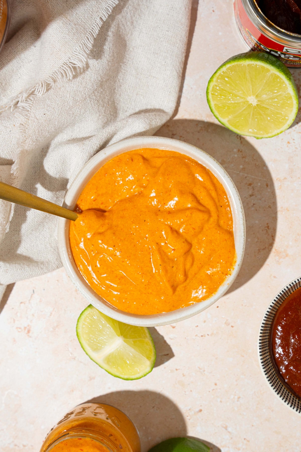 A white bowl with Taco Bell chipotle sauce with a spoon in the bowl. The bowl is on a tan counter with a jar of sauce, lime, and a white cloth napkin.