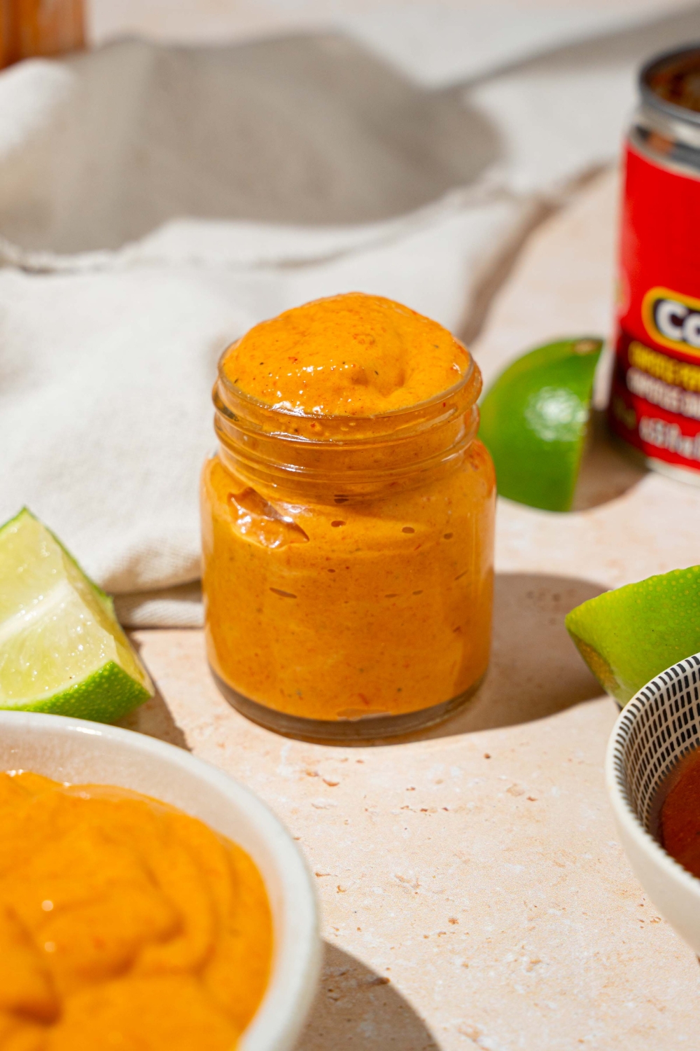A small glass jar with Taco Bell chipotle sauce. The jar is on a tan counter with a bowl of sauce, lime, and white cloth napkin.