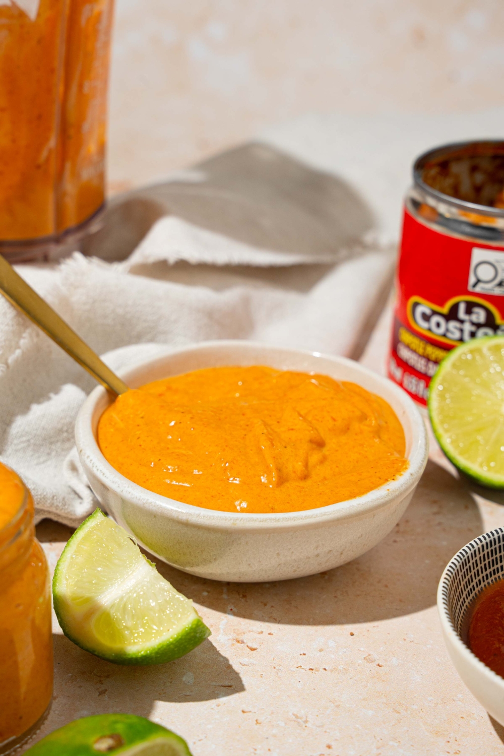 A white bowl with Taco Bell chipotle sauce with a spoon in the bowl. The bowl is on a tan counter with a jar of sauce, lime, and a white cloth napkin.