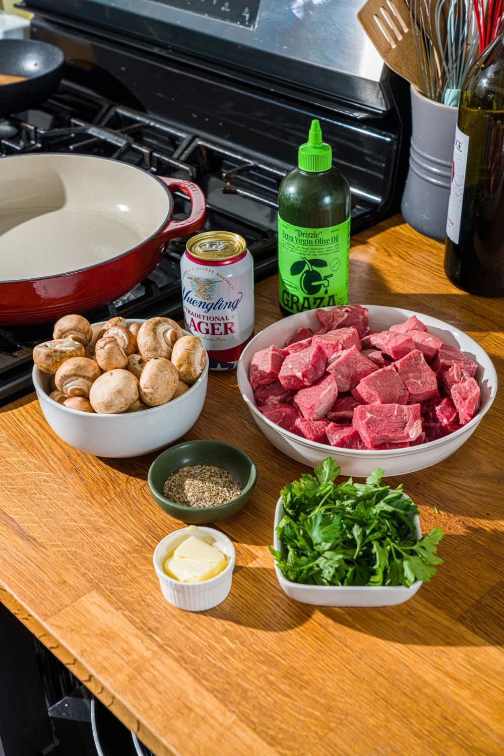 A wooden counter with ingredients to make tenderloin tips including beef tips, mushrooms, seasonings, oil, butter, and a can of beer.