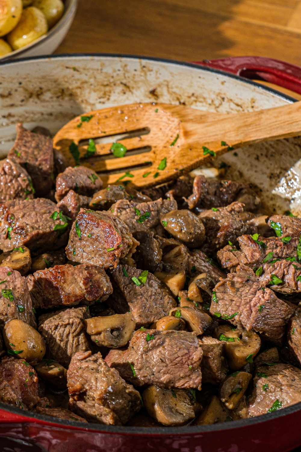 A cast iron skillet with tenderloin tips cooked with mushrooms and garnished with fresh parsley. There is a wooden spoon in the skillet.