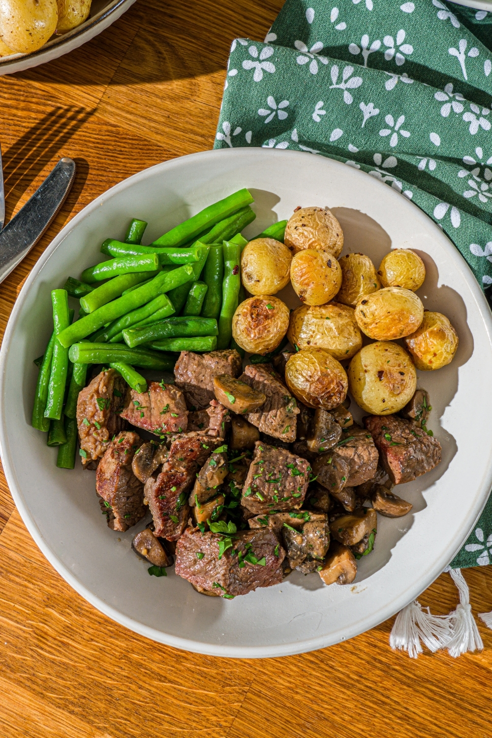 A white plate with tenderloin tips cooked with mushrooms and served with roasted potatoes and green beans. The plate is on a wooden counter with a green cloth napkin.