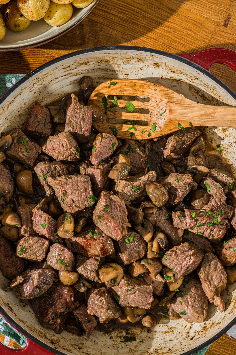 A cast iron skillet with tenderloin tips cooked with mushrooms and garnished with fresh parsley. There is a wooden spoon in the skillet.