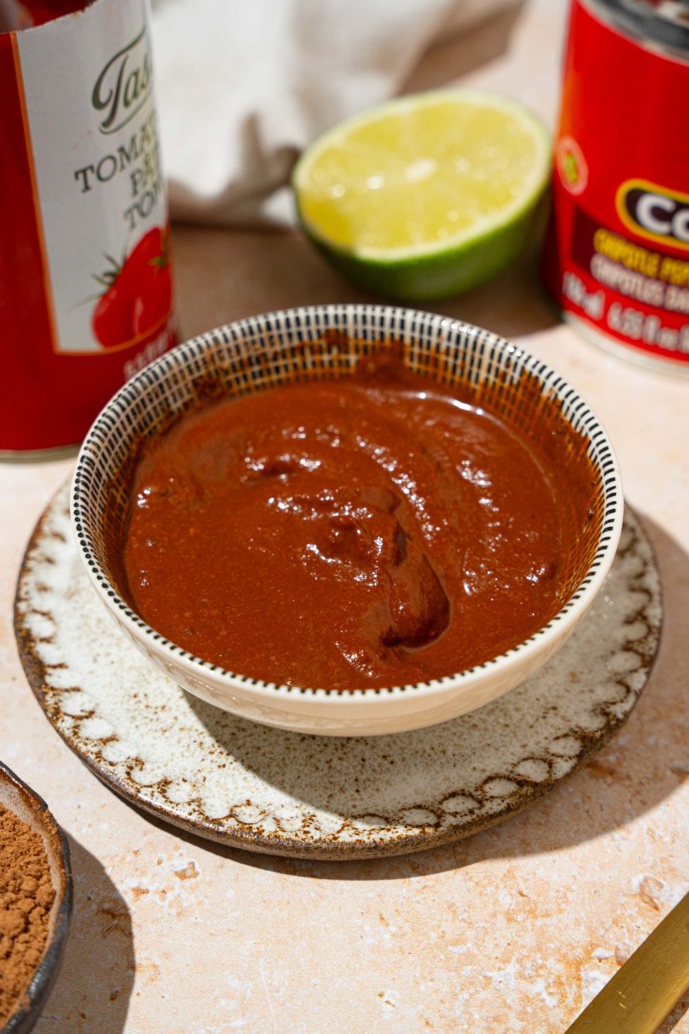 A white bowl of Tex Mex paste on a small plate. The plate is on a tan counter with a lime and cans of ingredients.