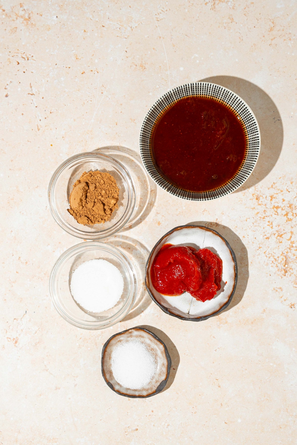 A tan counter with several bowls containing ingredients to make Tex Mex paste including abodo sauce, cocoa powder, tomato paste, salt, and sugar.