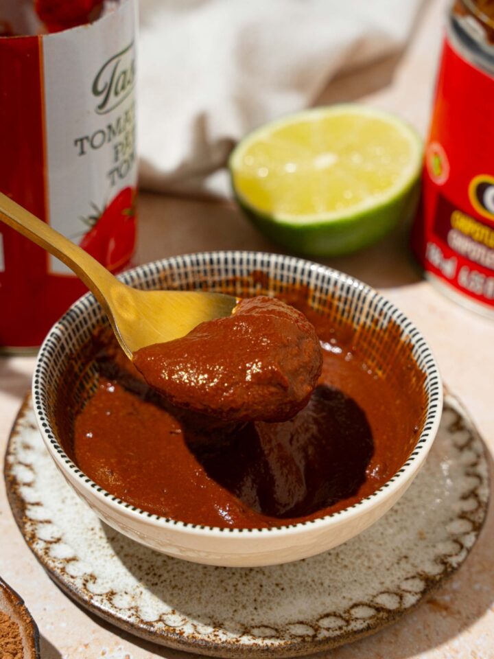 A white bowl of Tex Mex paste on a small plate. A spoon is dipping in the bowl. The plate is on a tan counter with a lime and cans of ingredients.