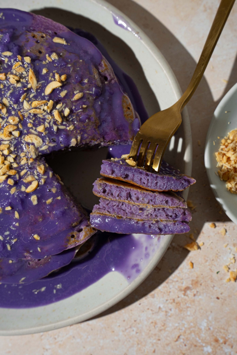 A white plate with a stack of ube pancakes topped with ube sauce and toasted coconut. A fork has taken a bite from the stack. The plate is on a tan counter.