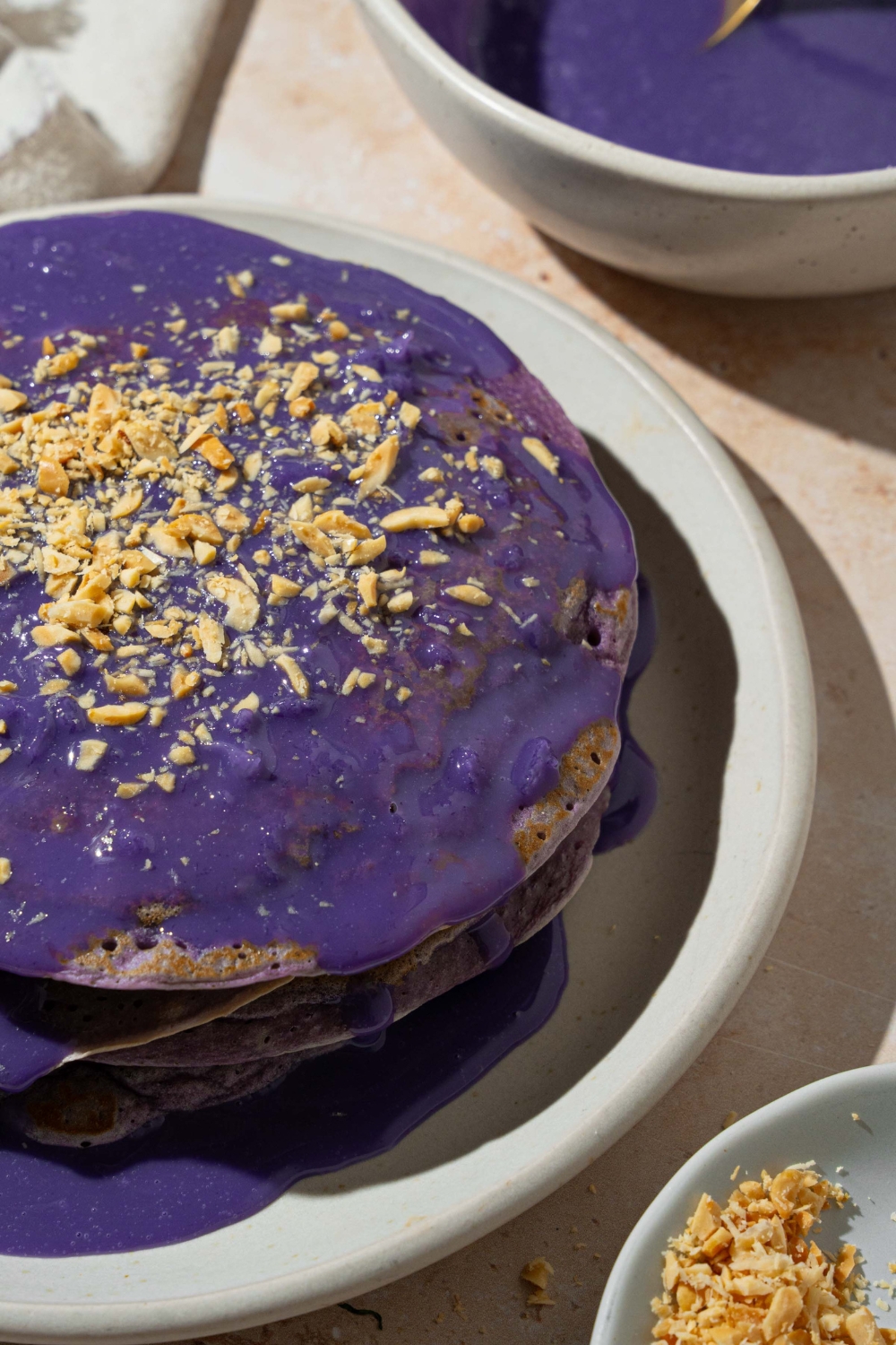 A white plate with a stack of ube pancakes topped with ube sauce and toasted coconut. The plate is on a tan counter with a white cloth napkin and bowl of ube sauce.