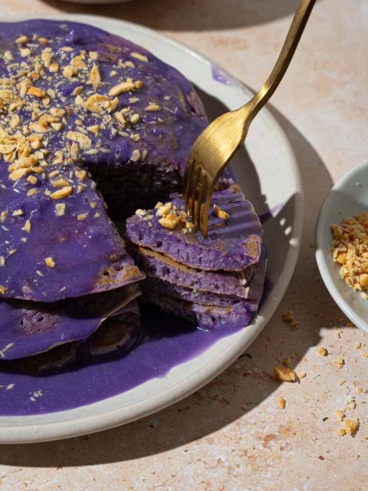 A white plate with a stack of ube pancakes topped with ube sauce and toasted coconut. A fork has taken a bite from the stack. The plate is on a tan counter.