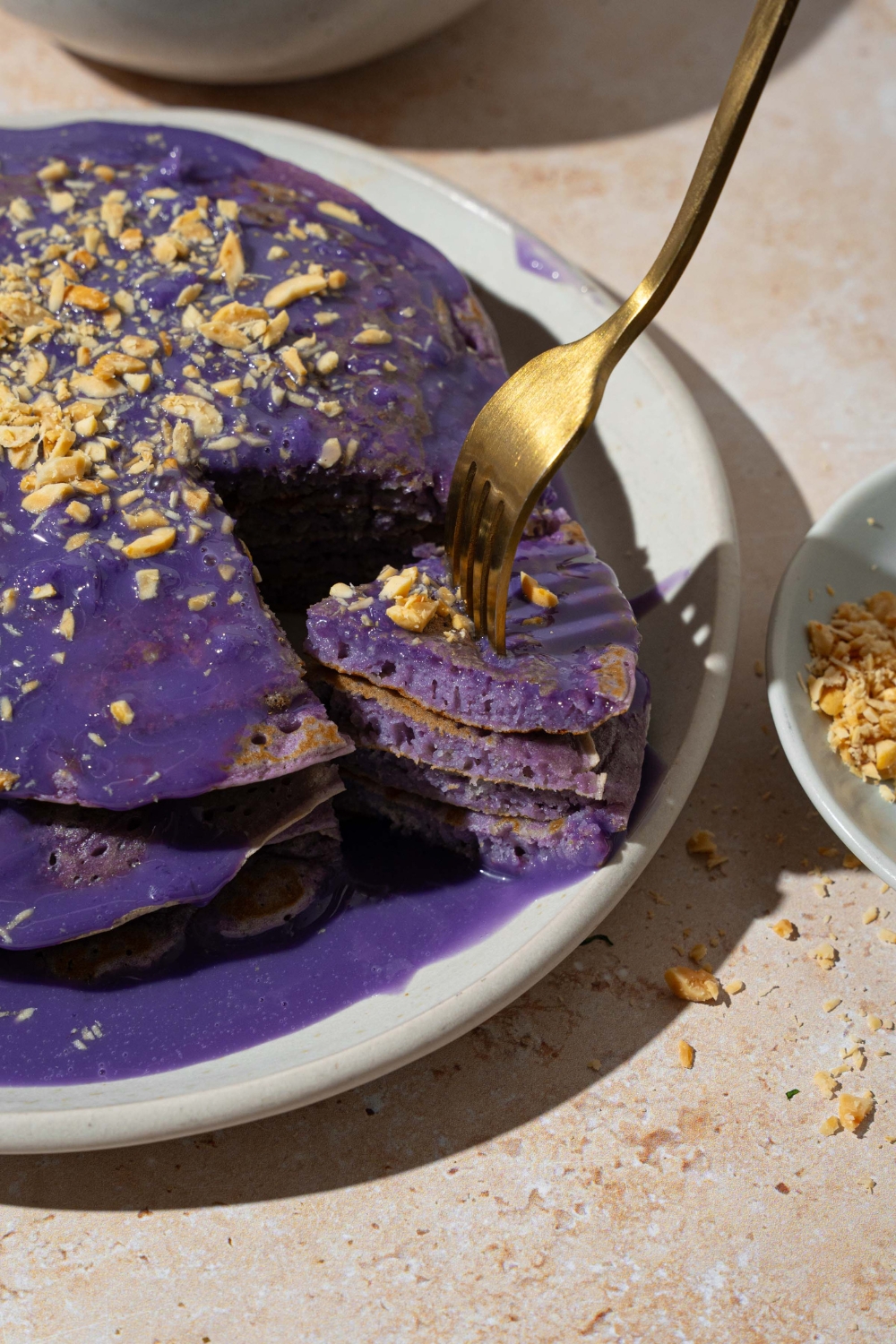 A white plate with a stack of ube pancakes topped with ube sauce and toasted coconut. A fork has taken a bite from the stack. The plate is on a tan counter.
