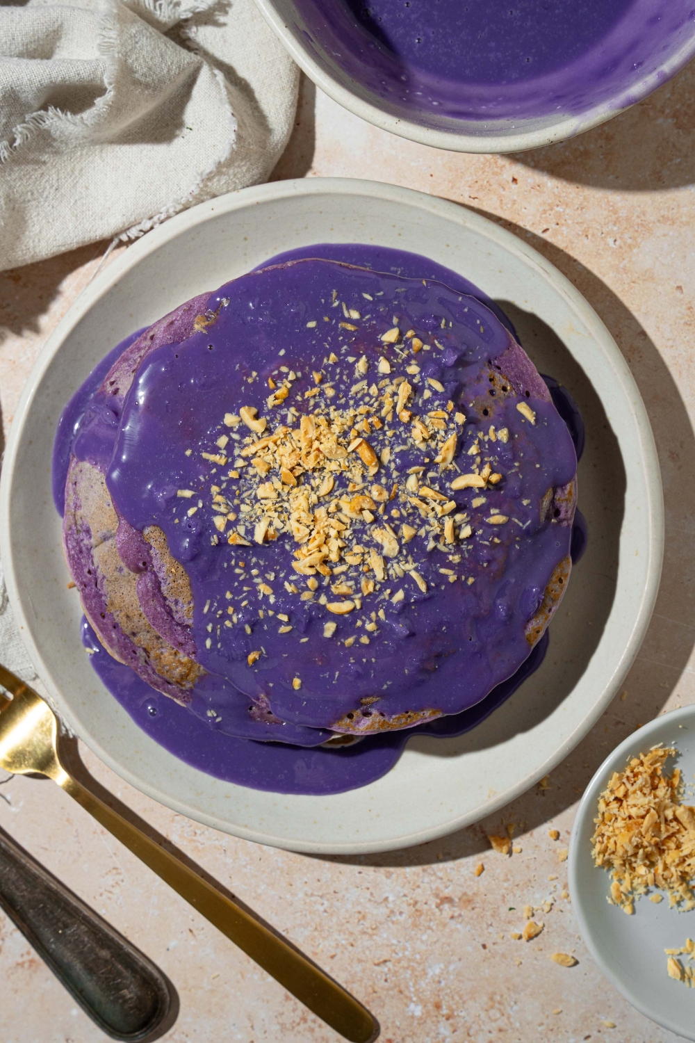 A white plate with a stack of ube pancakes topped with ube sauce and toasted coconut. The plate is on a tan counter with a white cloth napkin and bowl of ube sauce.