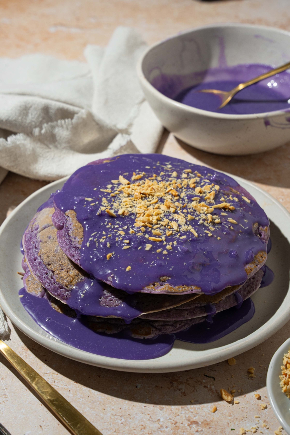 A white plate with a stack of ube pancakes topped with ube sauce and toasted coconut. The plate is on a tan counter with a white cloth napkin and bowl of ube sauce.