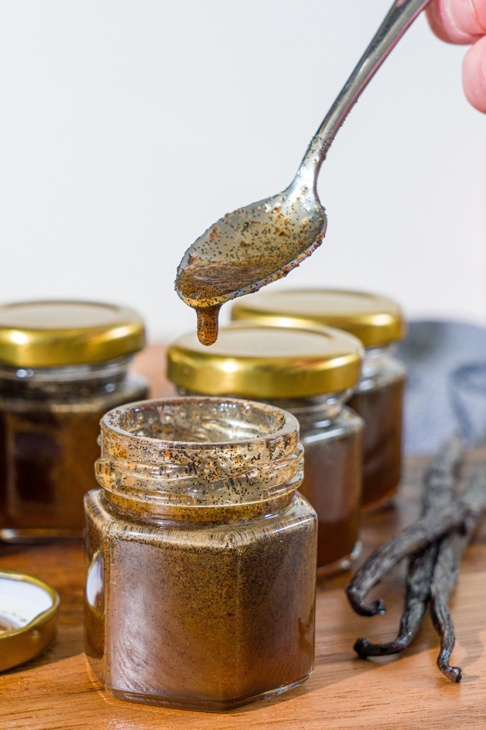 A wooden board with several jars of homemade vanilla bean paste and whole vanilla beans. A spoon is pouring the paste into one of the open jars.