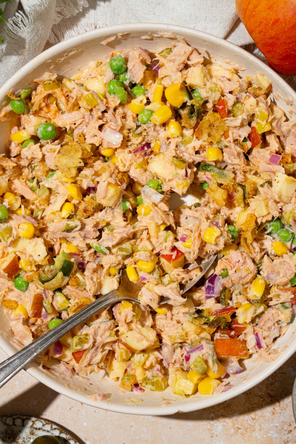 A large white bowl with Matthew McConaughey tuna salad including tuna, corn, peas, apple, pickles, red onion, and jalapeño chips in a wasabi mayo dressing. There is a spoon in the bowl. The bowl is on a tan counter with a white cloth napkin.