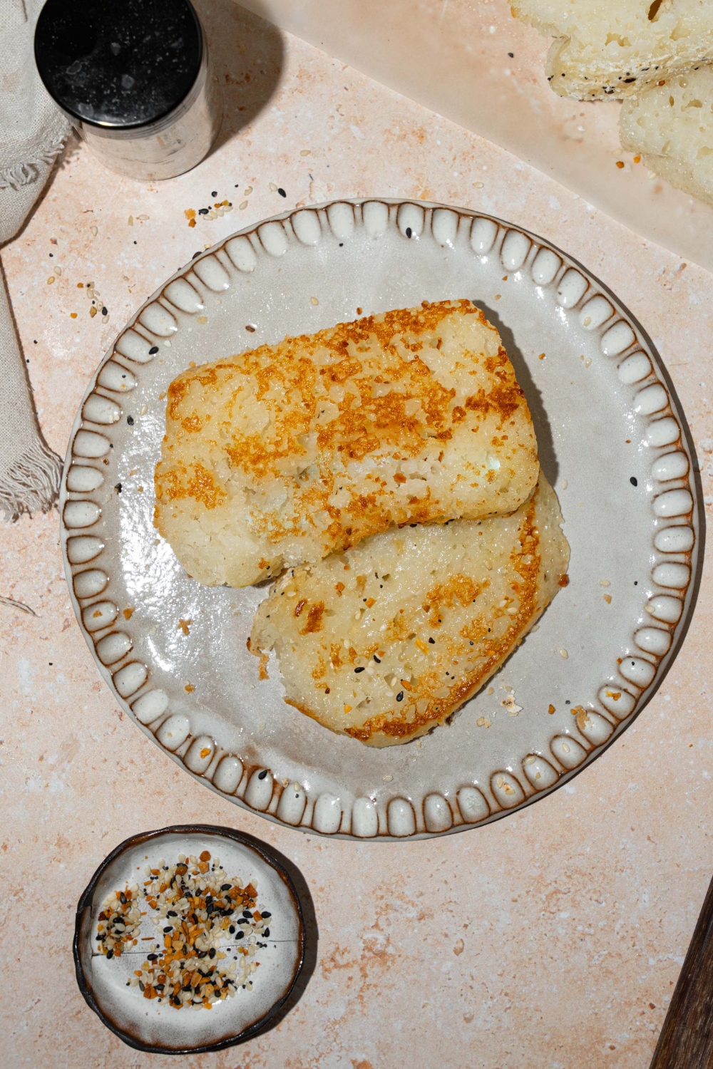A white plate with two slices of blender rice bread. The plate is on a tan counter with a loaf of sliced bread topped with everything bagel seeds.