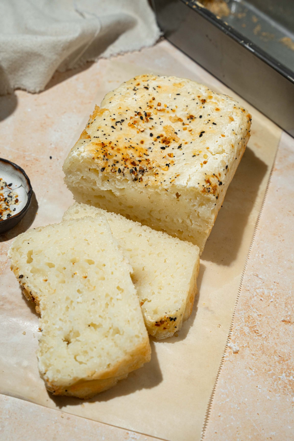 A loaf of sliced blender rice bread topped with everything bagel seeds. The loaf is on a piece of parchment paper on a tan counter with a small bowl of seeds and a bread knife.